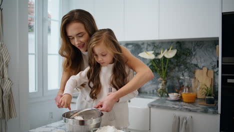 Adorable-child-helping-mom-at-kitchen-close-up.-Mother-daughter-sifting-flour