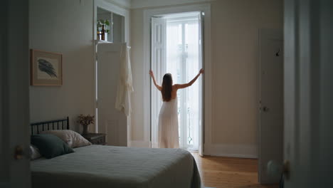 Young-woman-opening-blinds-home-morning.-Relaxed-girl-looking-window-back-view