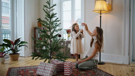 Positive-mom-daughter-putting-toys-fir-tree-indoors.-Pretty-girl-helping-mother