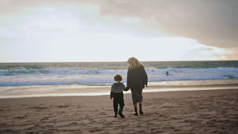 Mother-son-walking-beach-holding-hands-rear-view.-Carefree-family-resting-sunset