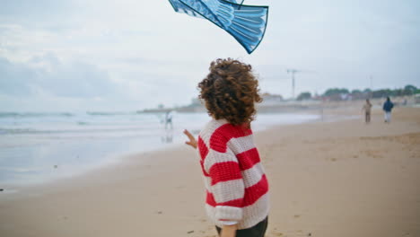 Little-boy-playing-kite-on-ocean-shore-rear-view.-Curly-kid-jumping-reaching-toy