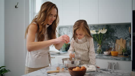 Smiling-mother-daughter-breaks-eggs-at-kitchen.-Child-mixing-ingredients-at-bowl