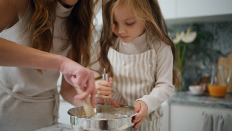 Focused-baby-mixing-whisk-at-kitchen-closeup.-Smiling-mother-helping-to-daughter