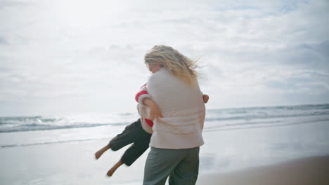 Playful-boy-running-mother-on-ocean-shore.-Happy-parent-woman-hug-spinning-child