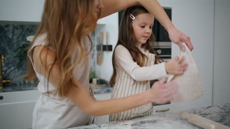 Focused-child-preparing-pastry-with-mom-at-home.-Positive-mother-holding-dough