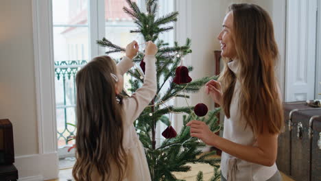 Lächelnde-Frau-Schmückt-Weihnachtsbaum-Mit-Mädchen-Zu-Hause.-Mutter-Hilft-Tochter