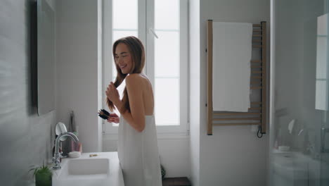 Positive-lady-arranging-hair-bathroom-interior.-Smiling-woman-holding-hairbrush