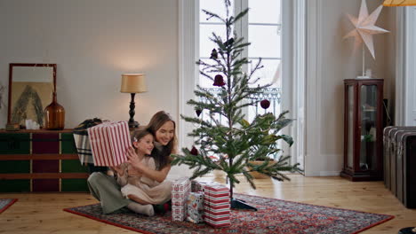 Süße-Familie-Schüttelt-Geschenkbox-In-Der-Nähe-Des-Weihnachtsbaums.-Frau-Umarmt-Mädchen-Im-Gemütlichen-Zuhause