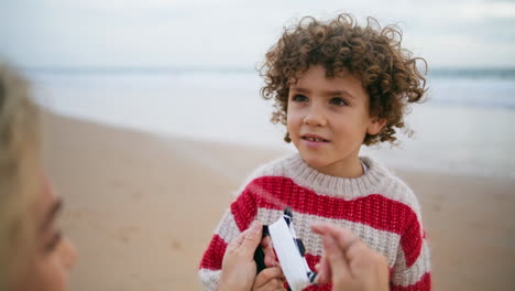 Portrait-adorable-curly-boy-resting-beach-with-mother.-Focused-child-hold-kite