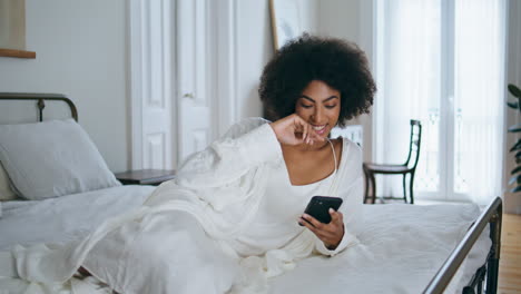 Laughing-model-watching-phone-funny-video-home.-Relaxed-african-lady-laying-bed