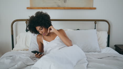 Awaking-girl-yawning-bed-at-morning.-African-woman-reading-phone-laying-pillow