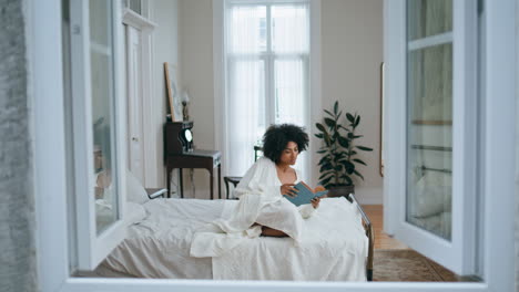 Carefree-woman-flipping-book-pages-indoor.-African-lady-reading-novel-laying-bed