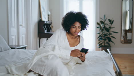 Smiling-lady-messaging-smartphone-at-bedroom.-Curly-woman-texting-sms-laying-bed