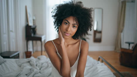 Gentle-model-sitting-bed-home-portrait.-African-woman-touching-dark-curly-hair