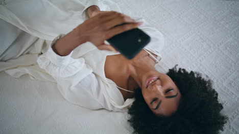 Serene-lady-reading-cellphone-resting-at-bed-top-view.-Curly-hair-woman-relaxing