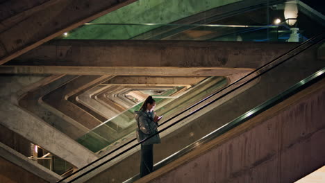 Girl-climbing-escalator-cellphone-at-dark-evening.-Trendy-woman-phone-on-stairs