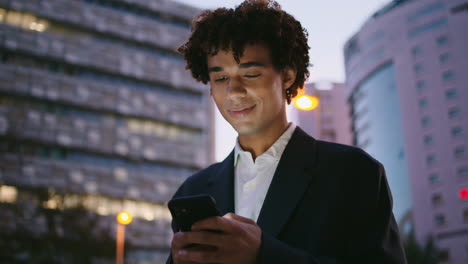Smiling-manager-browsing-smartphone-evening-downtown.-Curly-businessman-typing