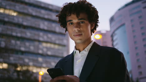 Formal-suit-guy-holding-phone-at-sunset-city-portrait.-Young-man-looking-camera