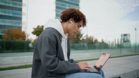 Curly-teenager-typing-laptop-at-street-closeup.-Young-man-listening-music-alone