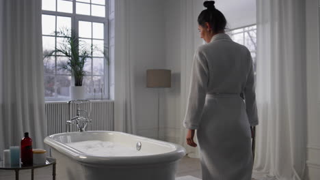 Bathrobe-model-entering-bath-room.-Calm-lady-touching-foam-preparing-spa-at-home