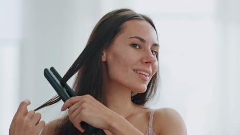 Happy-girl-using-hair-iron-at-white-bath-room-closeup.-Smiling-woman-hairstyle