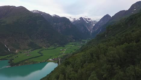 Melting-Briksdal-glacier-part-of-Jostedal-glacier-seen-from-lush-green-hillside-in-Olden-Norway---River-flowing-from-glacier-and-into-glacial-green-freshwater-lake-Oldevatn