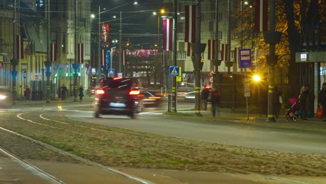 Timelapse-of-city-rush-hour-traffic-on-the-street-of-Liepaja,-traffic-light-streaks,-tram-rails-with-fast-moving-trams,-pedestrians-waiting-for-the-public-transport,-Latvian-flag,-distant-medium-shot
