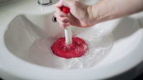 Red-and-white-plunger-unblocking-white-wash-hand-basin-sink,-mans-hand
