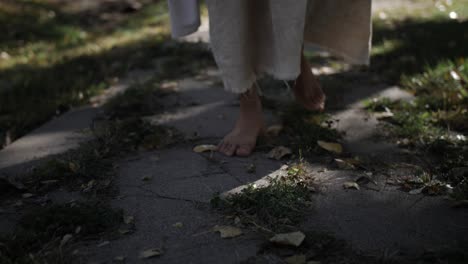 A-dramatic-slow-motion-closeup-of-Jesus'-feet-walking-through-grass-and-down-a-sidewalk-while-he-wears-a-white,-tattered-robe