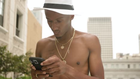 Close-up-of-a-man-wearing-fedora-hat-and-gold-chain-necklace-is-texting-or-looking-at-social-media-on-his-phone-beside-a-rooftop-swimming-pool