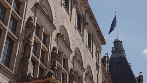 Panning-shot-of-Rotterdam-town-hall,-sandstone-building-with-statues-above-the-entrance-with-a-European-flag-flying