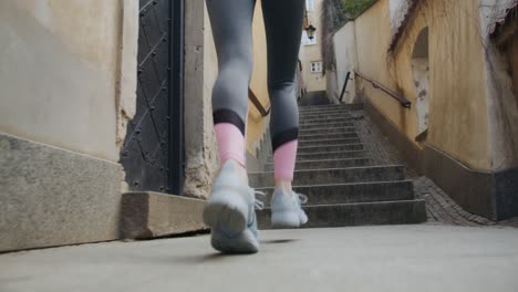Woman-jogging-and-running-up-stairs-in-an-alley-at-an-historic-town,-low-angle-tracking-shot-seen-from-behind