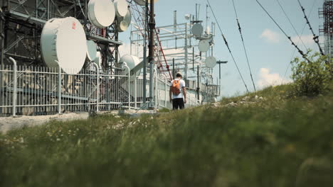 Hiker-with-a-cap-and-an-orange-backpack-walking-past-the-radio-tower-near-the-fence-away-from-the-camera