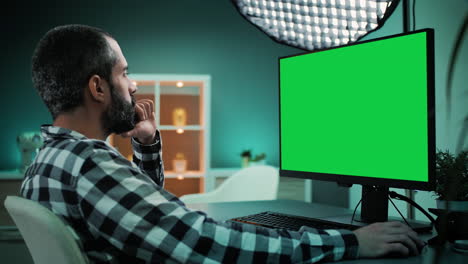 Slow-motion-shot-over-the-shoulder-of-a-handsome-and-focused-young-freelancer,-copywriter,-designer,-editor-or-content-creator-working-on-desktop-computer-with-chroma-key-green-screen-mock-up-display