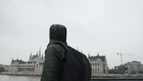 Boy-standing-in-front-of-Budapest-Parliament-from-the-river-side,-with-a-boat-on-the-front,-filmed-from-a-boat