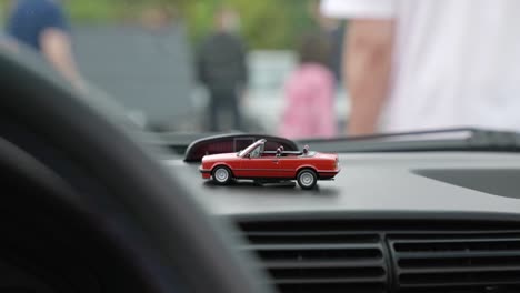 Tiny-red-toy-car-displayed-on-vehicle-dashboard-interior-as-people-walk-outside