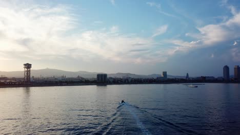 Ziehen-Sie-Die-Luftaufnahme-Eines-Bootes-Ein,-Das-Die-Küste-Von-Barcelona-Während-Eines-Wunderschönen-Sonnenuntergangs-Mit-Blick-Auf-Die-Skyline-Der-Stadt-Erreicht