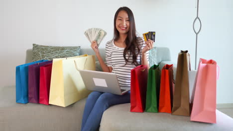 Satisfied-and-happy-looking-attractive-woman-sitting-an-a-couch-wearing-a-striped-shirt-and-blue-jeans-smiling-with-money-in-one-hand-and-credit-cards-in-the-other-surrounded-by-colorful-shopping-bags