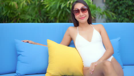 Young-attractive-adult-Asian-woman-in-white-swim-suit-and-sunglasses-sitting-on-bright-blue-outdoor-sofa-with-yellow-contrast-pillow-on-tropical-poolside-background