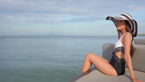 Una-Mujer-Asiática-Con-Traje-De-Baño-Y-Sombrero-Disfruta-Del-Sol-Al-Aire-Libre-En-Un-Resort-De-Playa