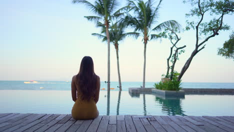 A-woman-with-her-back-to-the-camera-sits-on-a-wooden-jetty-while-looking-out-on-the-resort-infinity-pool-and-ocean-horizon