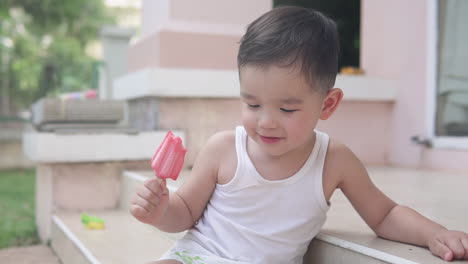 Slow-motion-video-of-an-Asian-toddler-boy-wearing-summer-clothes-sitting-on-the-stairs-outside-a-house-holding-a-red-strawberry-popsicle-ice-cream-smiling-and-thinking