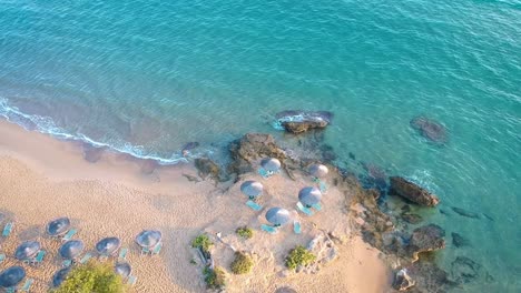 Aerial-view-over-the-crystal-clear-blue-water-reaching-the-sandy-beach-with-shacks-and-recliners-at-tourist-destination-in-Greece