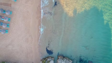 Drone-Mirando-Hacia-Abajo-Sobre-El-Agua-De-Mar-Tranquila-Que-Se-Encuentra-Con-La-Playa-De-Arena-Con-Sillones-Reclinables-Dispuestos-Horizontalmente-En-El-Destino-Turístico