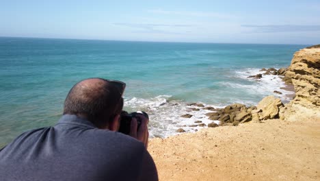 Man-takes-picture-or-video-of-seagull-flying-over-rocky-ocean-cove,-Slow-Motion