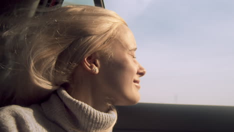 Woman-smiles-out-open-car-window-as-wind-blows-long-blond-hair,-close-up