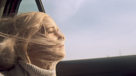 Woman-looks-out-open-car-window-as-blond-hair-swirls-across-face,-close-up
