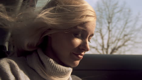 Young-woman-smiles-as-wind-blows-long-blond-hair-in-car-window,-close-up
