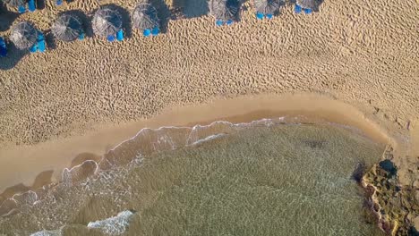 Aerial-Pedestal-Over-Waves-Crashing-On-Beach-With-Empty-Sunbeds-With-Blue-Parasols