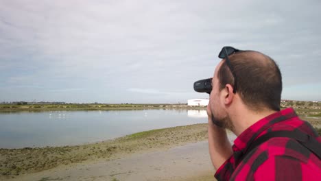 Man-Bird-Watching-Flamingos-with-Binoculars,-Over-the-Shoulder-Closeup